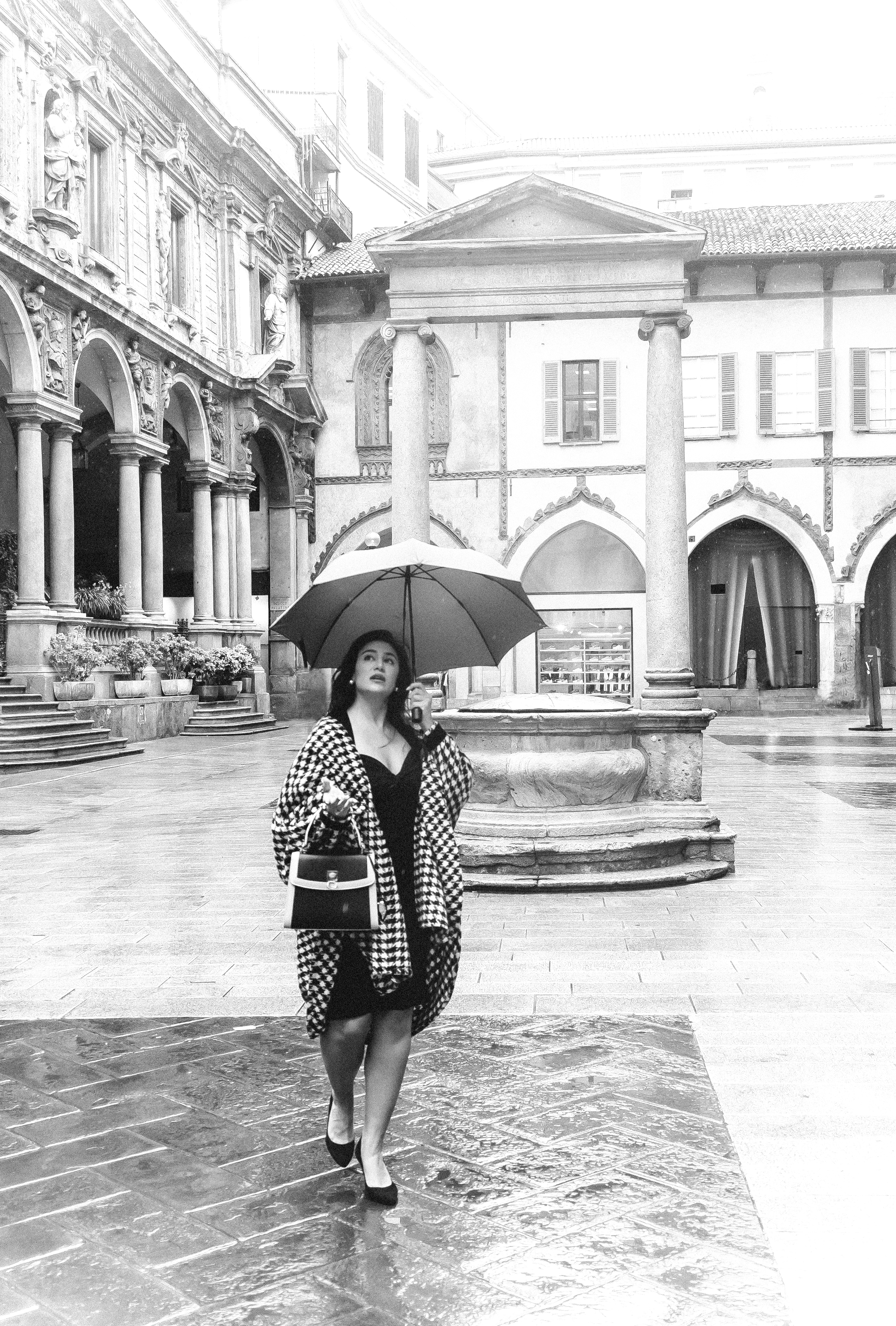 Italian woman walking in the rain on a piazza, gracefully holding an umbrella and an ILNI handbag.